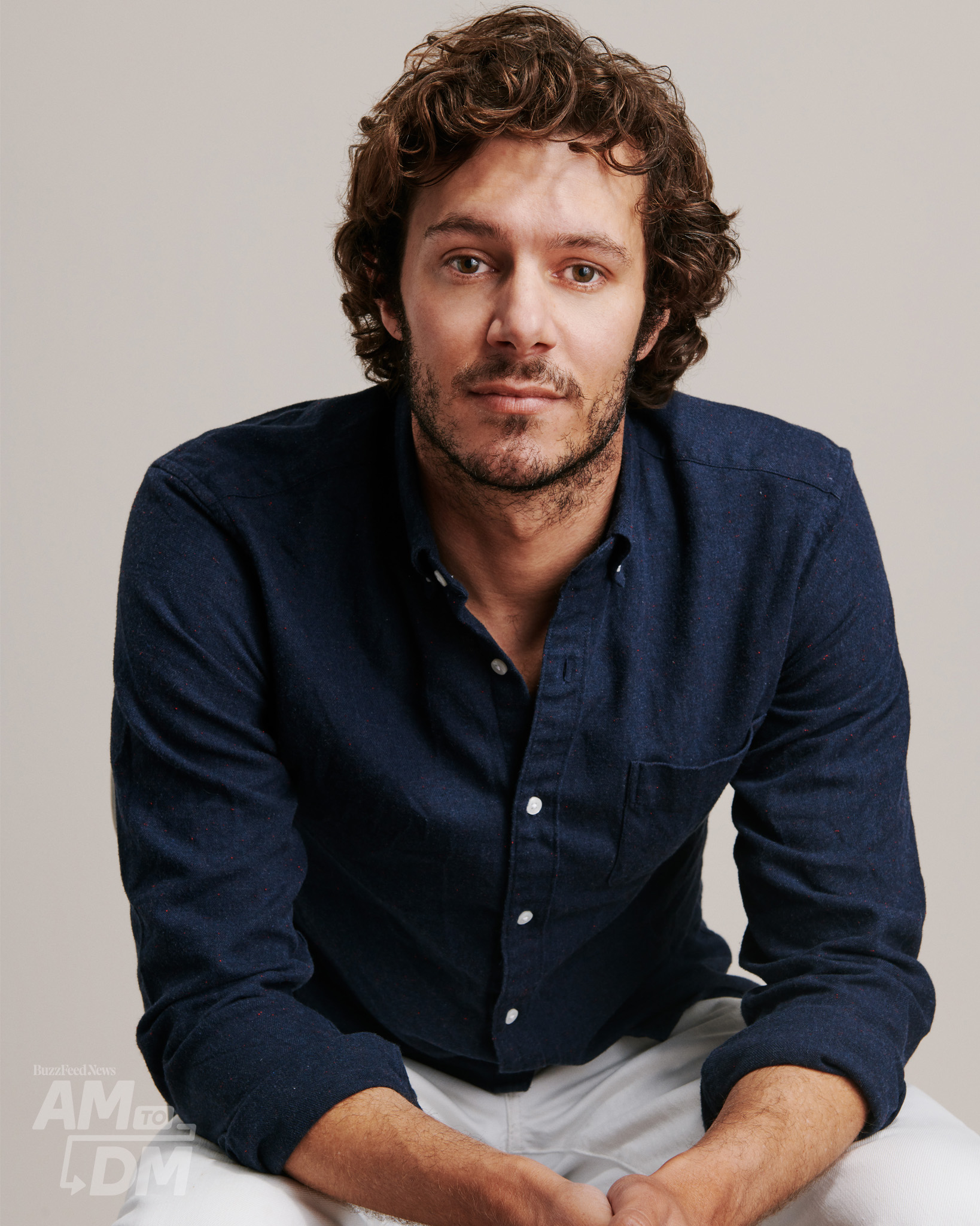Portrait of Adam Brody wearing a dark blue shirt and white pants in front of a taupe background CREDIT: JJ Reddington / BuzzFeed News