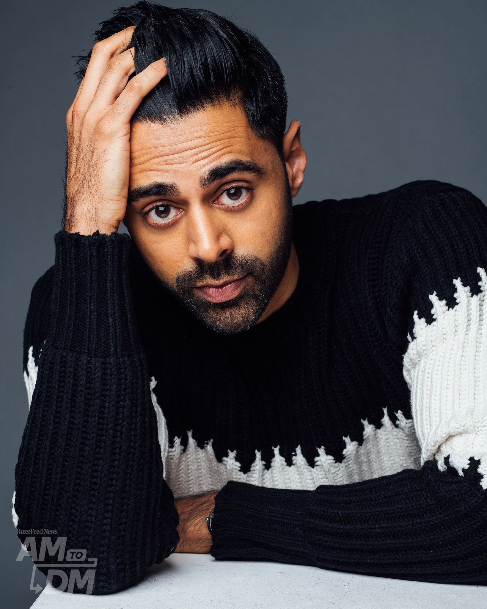 Portrait of Hasan Minhaj wearing a black and white sweater in front of a gray background. CREDIT: Taylor Miller / BuzzFeed News
