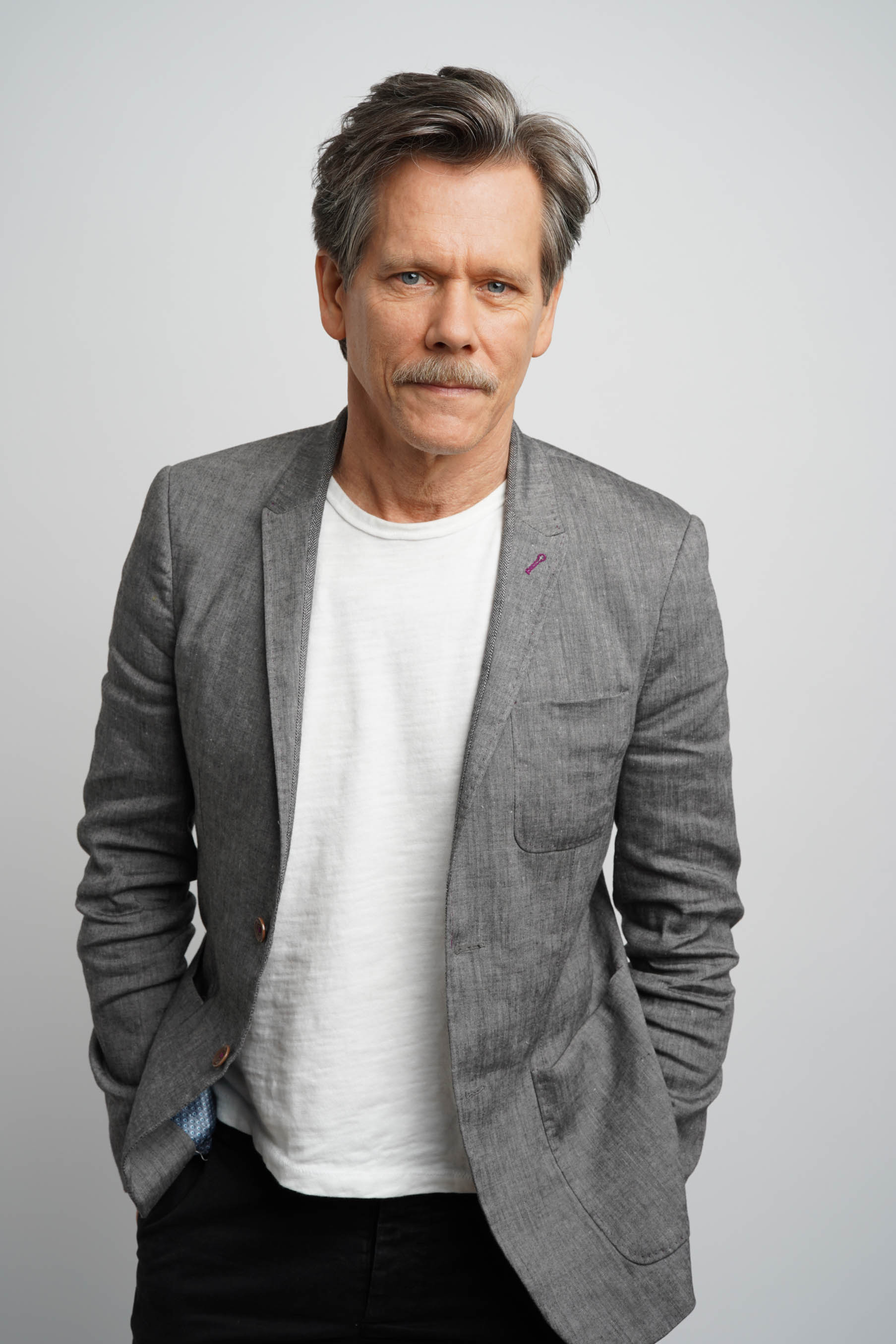 Portrait of Kevin Bacon wearing a gray blazer over a white t-shirt in front of a light gray background. CREDIT: Erich Bartlebaugh / BuzzFeed News