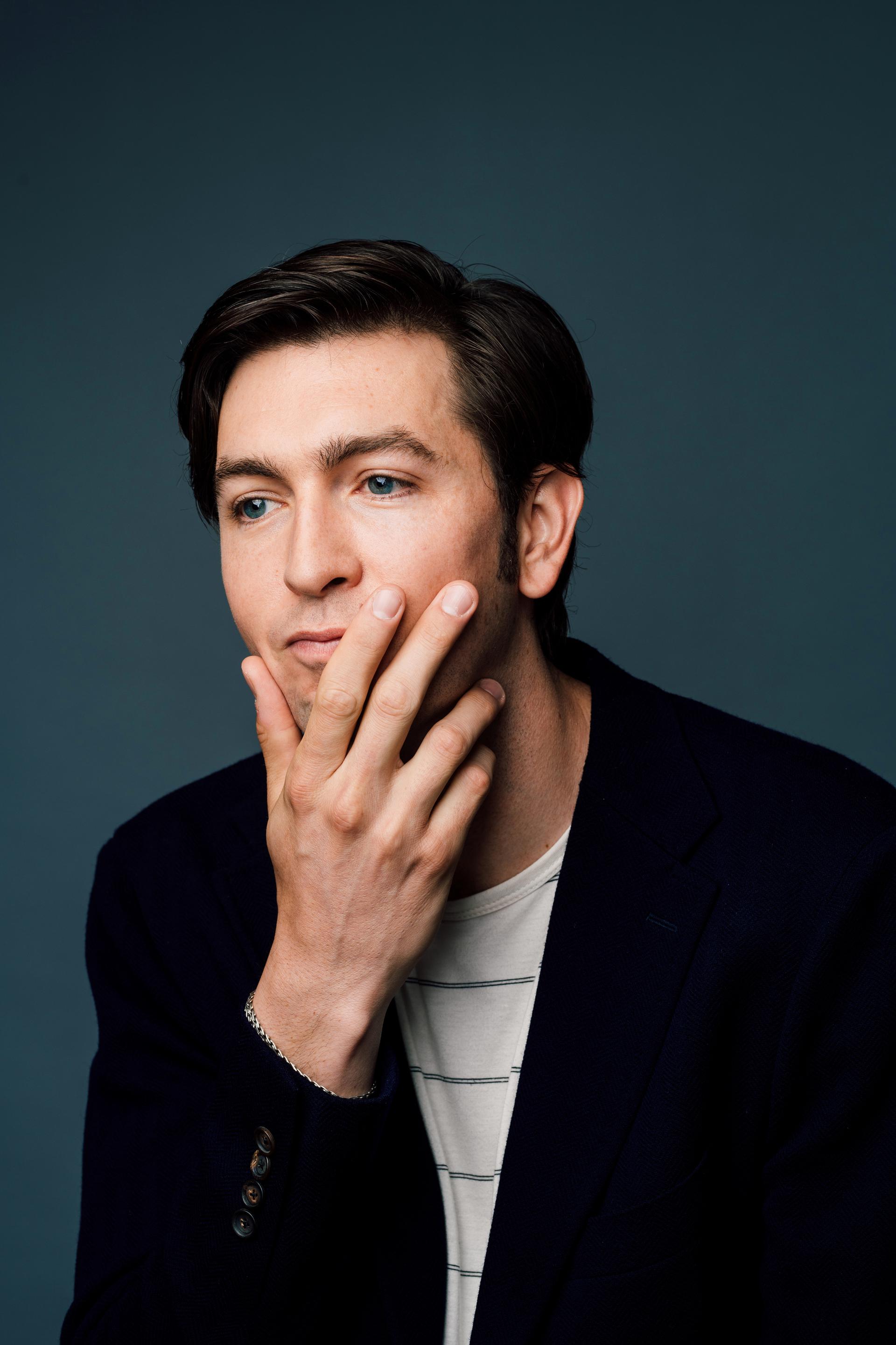 Portrait of Nicholas Braun wearing a black blazer over a striped t-shirt in front of a dark gray background. CREDIT: Taylor Miller / BuzzFeed News
