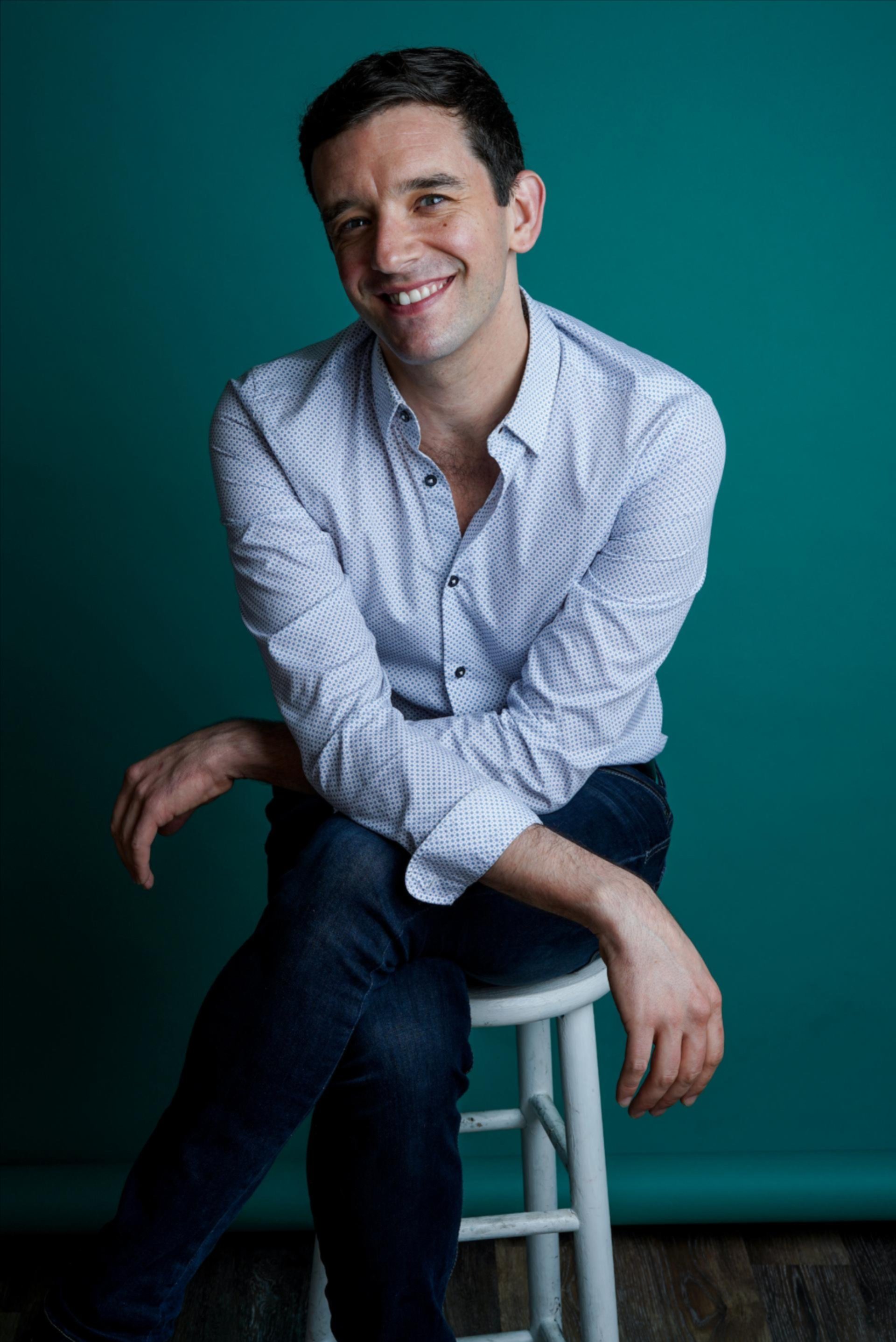 Portrait of Michael Urie wearing a light blue button down shirt over a white t-shirt sitting in front of a light green background. CREDIT: Erich Bartlebaugh / BuzzFeed News